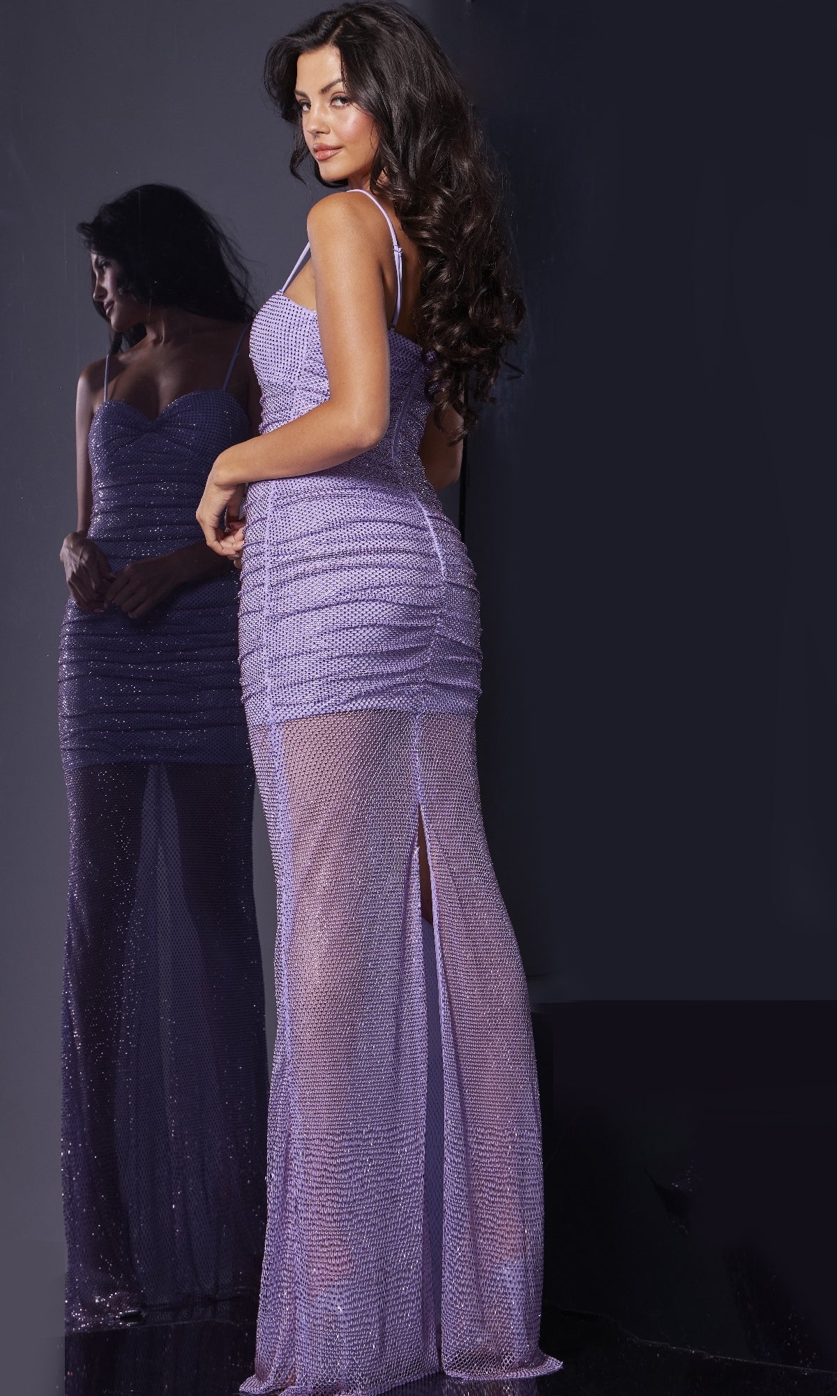 A woman wears the Jovani 38188 Long Prom Dress, a fitted, sleeveless lavender gown with sparkles, high slit, and sweetheart neckline, standing by a mirror and facing slightly away from the camera.